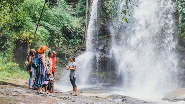Group of travelers listening to a guide near a cascading waterfall in a lush jungle setting.