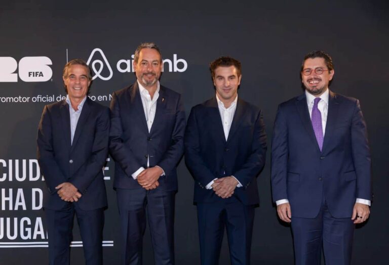 Four men in dark suits stand in a row posing for a photo against a dark backdrop with logos visible above them (FIFA and Airbnb). The group appears at a formal event or press moment, all smiling.