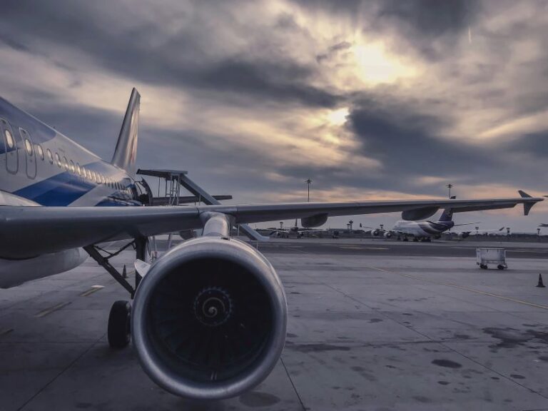 Front view of a jet engine and wing on the tarmac at an airport, with distant planes under a cloudy sunset sky.