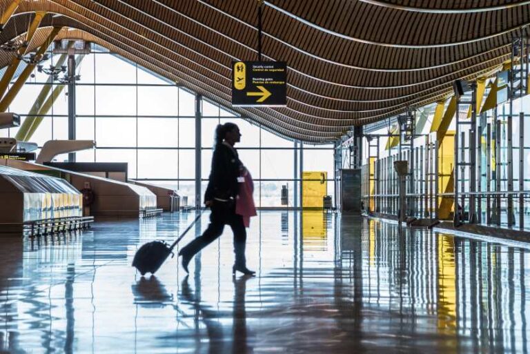 Traveler pulling a rolling suitcase through a spacious, sunlit airport terminal with a curved wooden ceiling and glass walls.