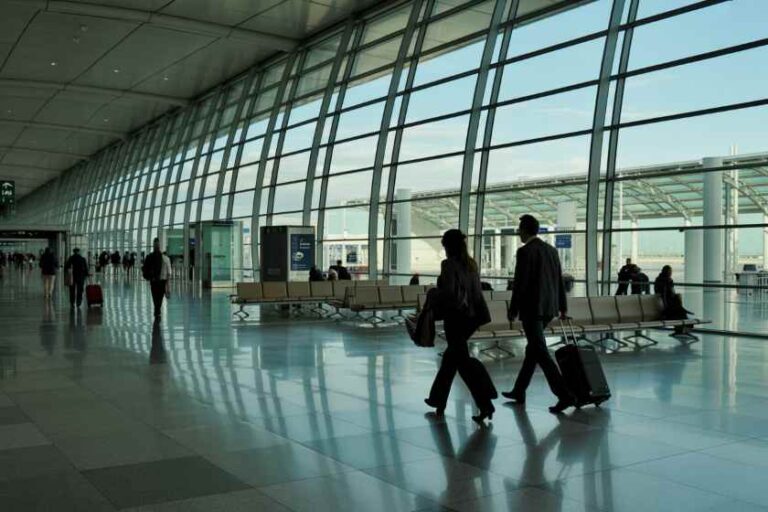 Airport terminal with floor-to-ceiling glass walls; travelers walk with rolling suitcases in bright, spacious concourse.
