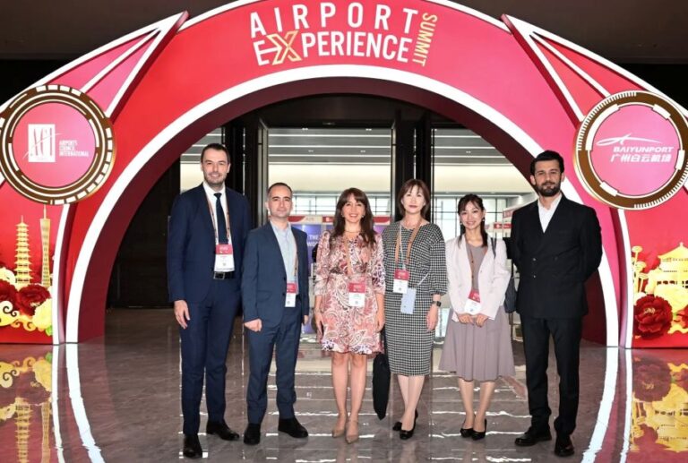 Six professionals in business attire pose for a group photo under a pink arch labeled Airports Experience Summit at the venue entrance.