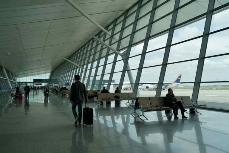 Airport terminal interior with large glass windows; traveler walks with rolling suitcase while others sit in rows of chairs.