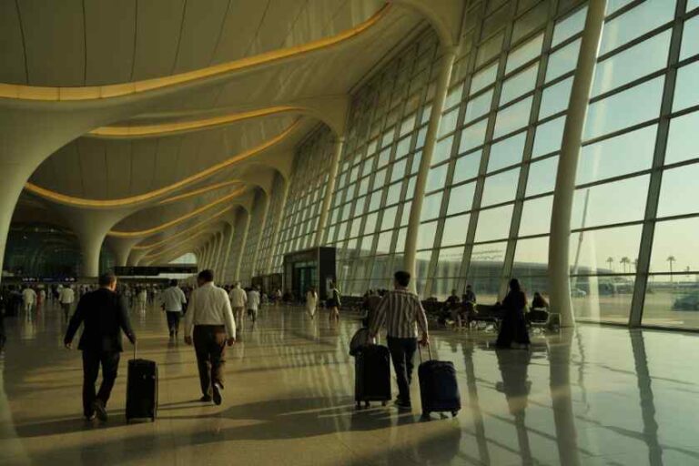 Travelers with rolling suitcases walk through a bright, modern airport concourse with large curved ceilings and expansive windows.