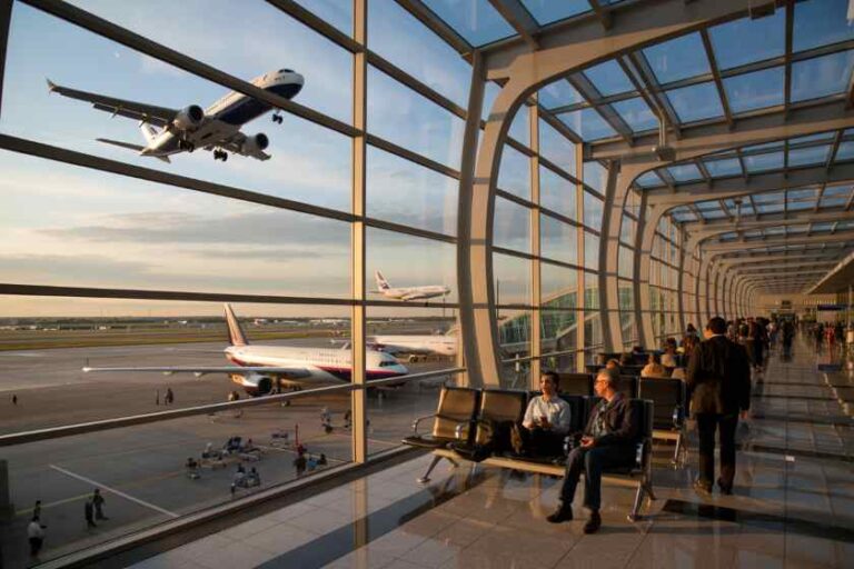 Passengers seated in a glass-walled airport terminal as planes taxi and take off on the runway outside at sunset.