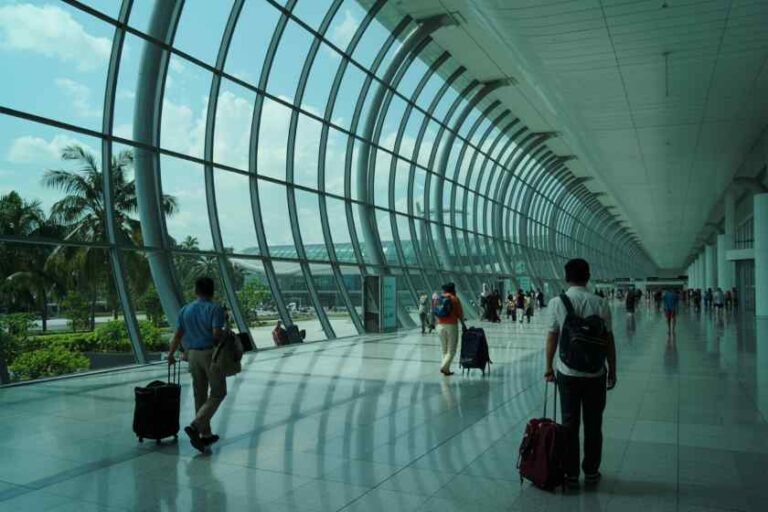 Wide, modern airport terminal with curved glass walls and travelers pulling suitcases along a sunny interior corridor.