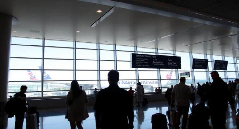 Busy airport terminal with travelers standing and sitting near large windows; overhead signs point to gates and services outside view of jet.