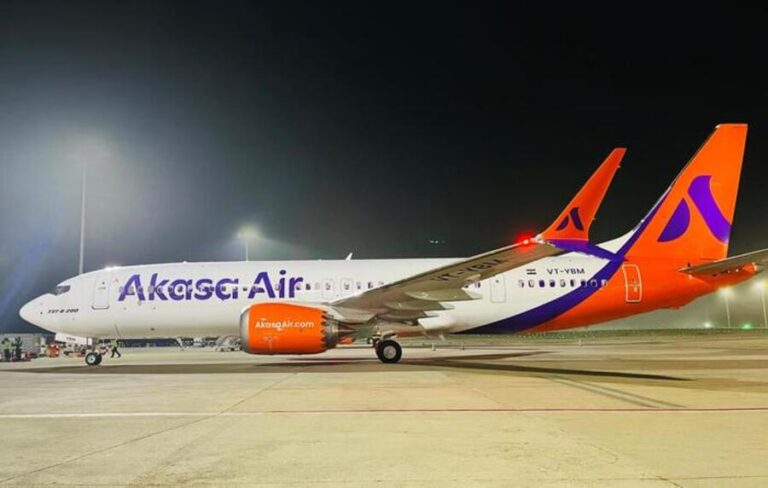 A Akasa Air passenger aircraft parked on the runway at night, white fuselage with purple Akasa Air branding and orange engine, orange tail with purple logo under bright field lights