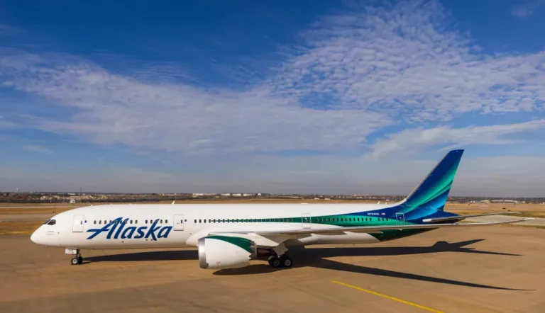 Side view of an Alaska Airlines passenger jet parked on a taxiway under a clear blue sky, with teal-green tail branding visible.