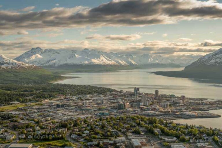 Aerial view of a coastal city by a calm bay with snow-capped mountains in the distance.