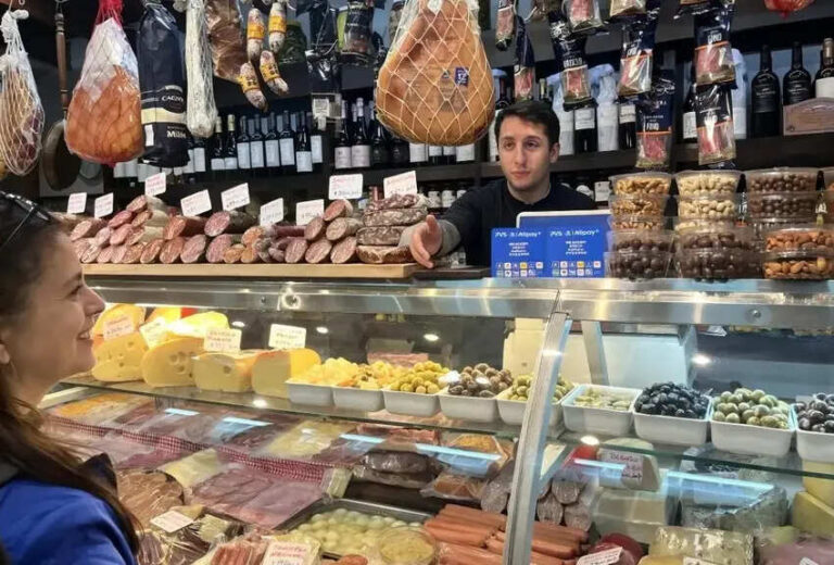 Smiling customer at a deli counter with cheeses, olives, and sliced meats behind glass, while a shopkeeper assists from the back wall.