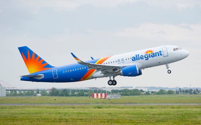 Allegiant Air jet lifting off from a runway with green fields in the foreground and a blue-orange livery.