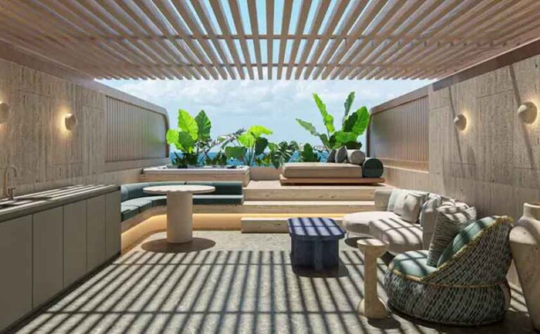 Outdoor rooftop lounge with seating, plants, and striped shade casting shadows on the floor, against a blue sky.