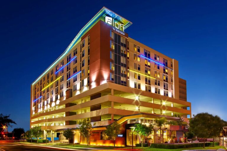 Nighttime exterior of a modern multi-story hotel with colorful neon lights along the facade and a parking podium below.