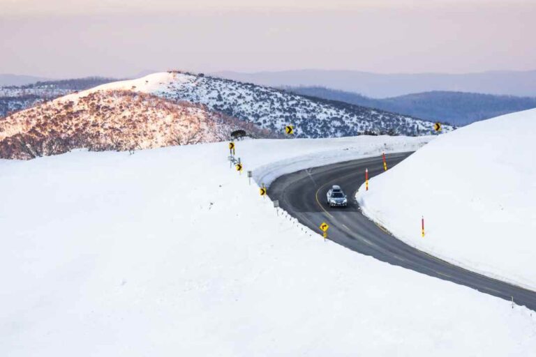 Car driving along a winding snow-covered mountain road with yellow curve signs and plowed asphalt.