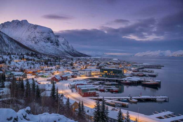 Snowy harbor town at dusk, with lit houses, boats docked along icy piers and snow-covered mountains in the background.