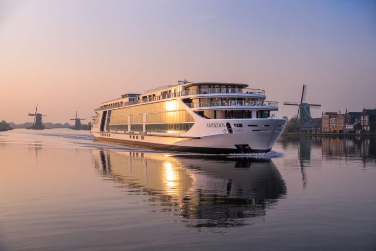 White cruise ship sailing on calm water at sunset with windmills and a village in the background.