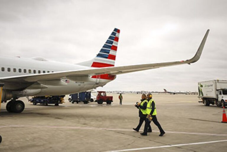 Airport ground crew in high-visibility vests walk beside a large commercial airplane on the tarmac by its wing and tail with American Airlines livery.