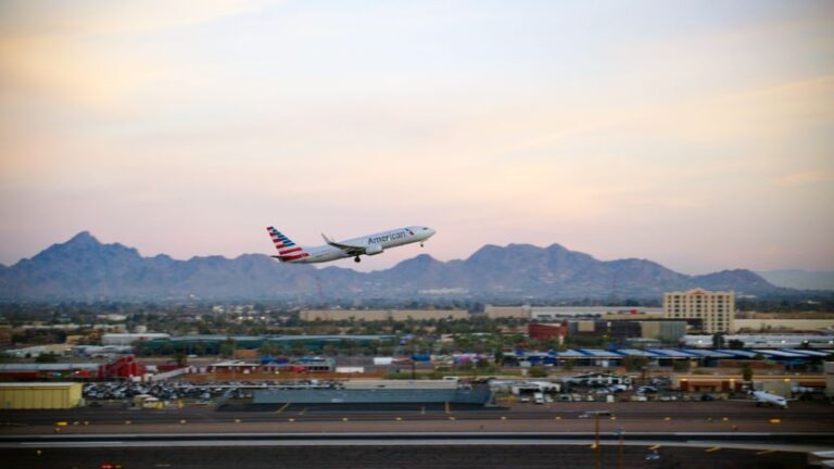 American Airlines jet taking off from a runway at sunset with desert city and distant mountains in the background.