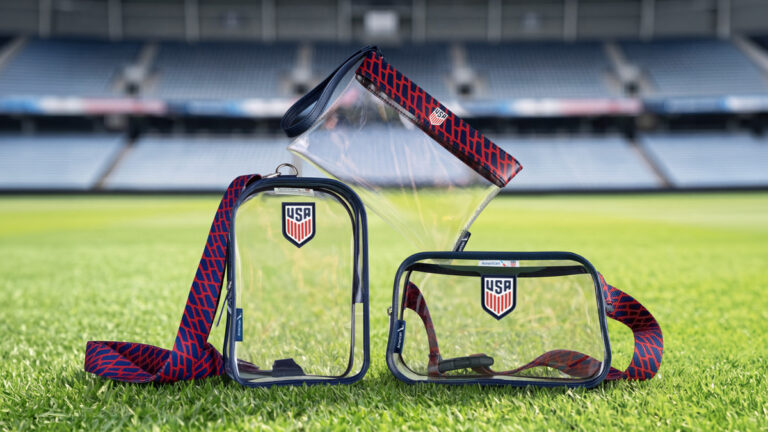 Three clear USA-branded stadium bags with red-and-navy patterned straps on a grass field.