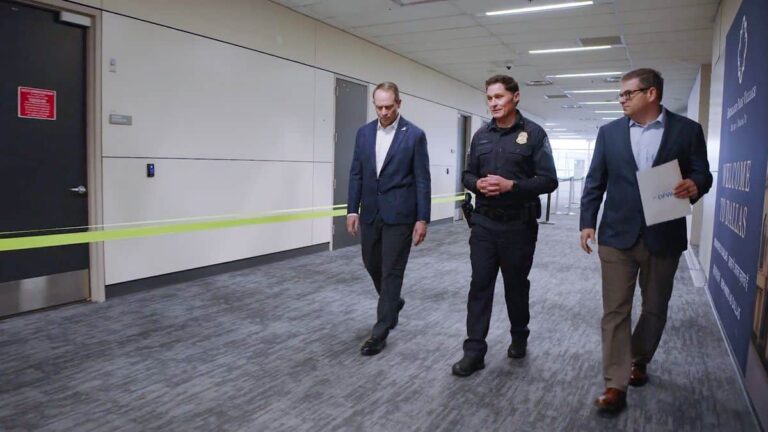 Three men walking a corporate hallway past yellow tape barrier; one in a police uniform, two in suits.