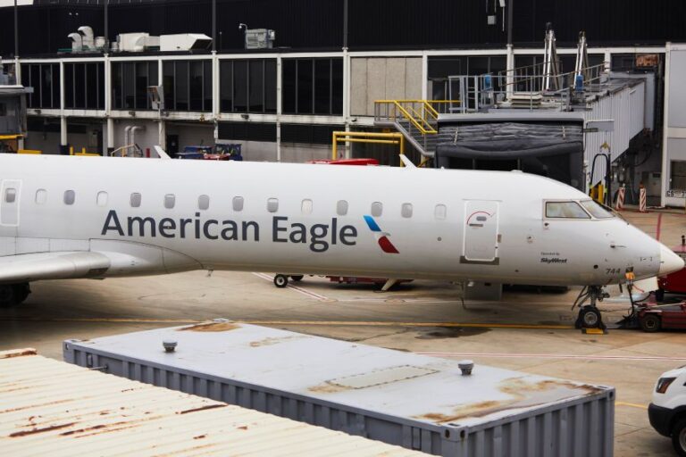 White American Eagle regional jet parked at a gate on the tarmac, with a jet bridge and airport terminal visible behind it.