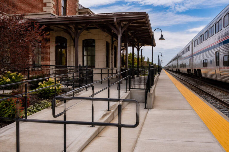 Train station platform with a long silver train on the right and a brick station building on the left under a blue sky, daylight.13?