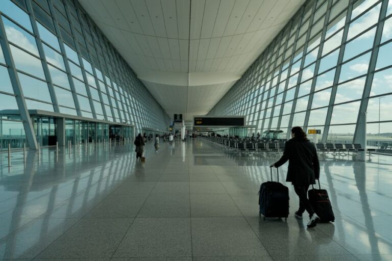 A person pushing a suitcase cart through a spacious airport terminal with glass walls and bright natural light.