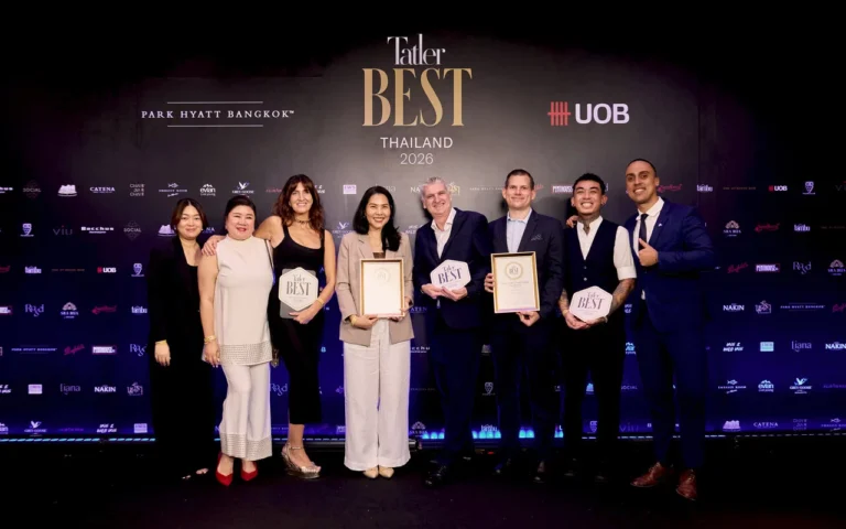 Eight people in formal wear pose on a dark stage at the Tatler Best Thailand 2026 event, some holding framed plaques and certificates with a sponsor backdrop behind them.