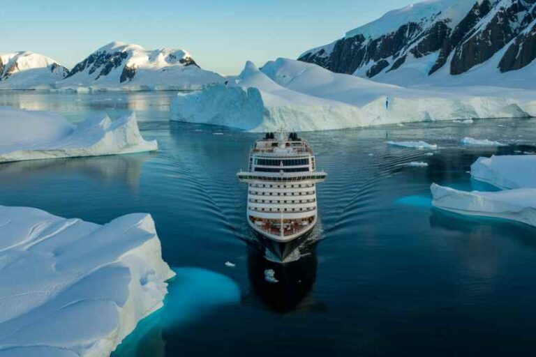 Cruise ship sailing through icy blue waters with towering icebergs and snow‑capped mountains in the background.