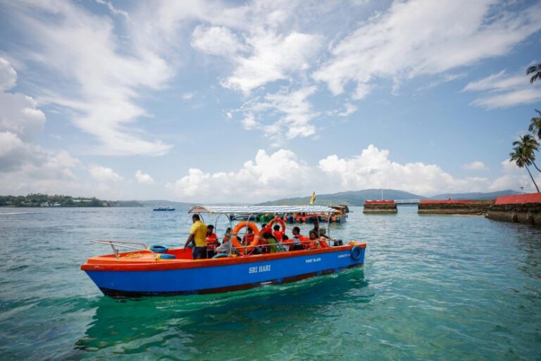 Blue and orange passenger boat with people wearing life jackets cruising in clear tropical water near a coastline.