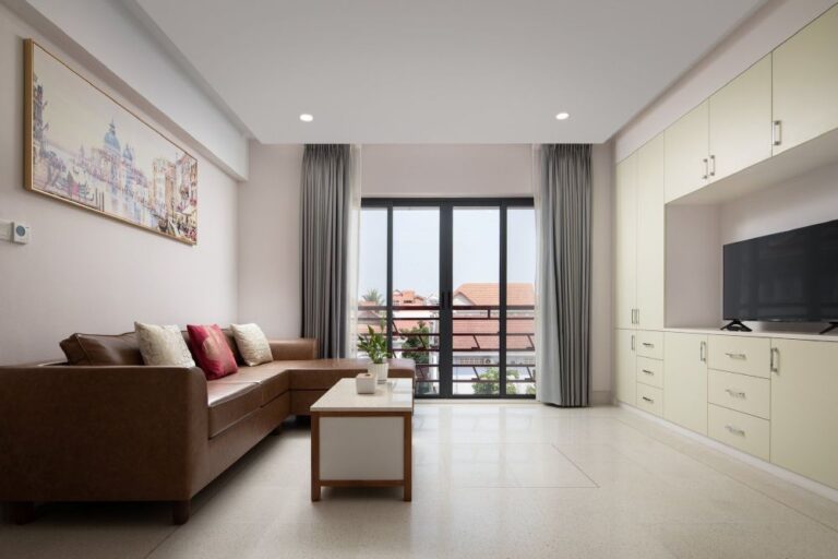 Bright living room with a brown sectional sofa, coffee table, and built-in cabinets with a TV, large window with gray curtains, overlooking rooftops.