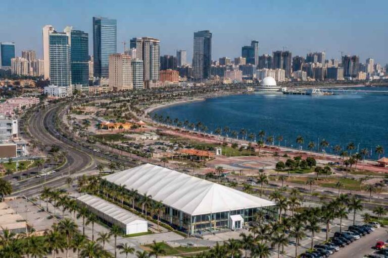 City skyline with a curved coastal road, palm trees, and a large white tent-like pavilion in the foreground by the water.