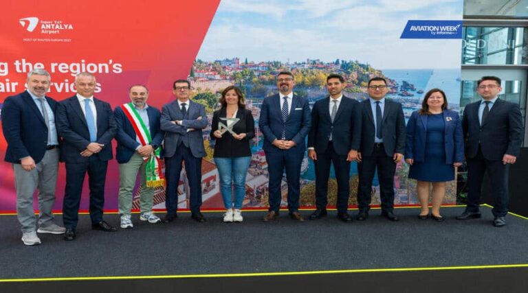 Nine professionals in business attire stand in a line on a stage with a red backdrop and coastal city backdrop, one man wearing an Italian sash and others holding a trophy or posing for a photo.