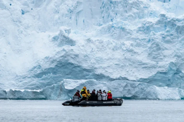 Group of people in life jackets aboard a rigid inflatable boat near a towering blue iceberg.