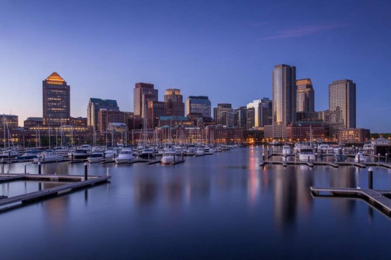 Blue hour: city skyline with tall buildings reflected on calm harbor, numerous sailboats moored at docks in foreground.