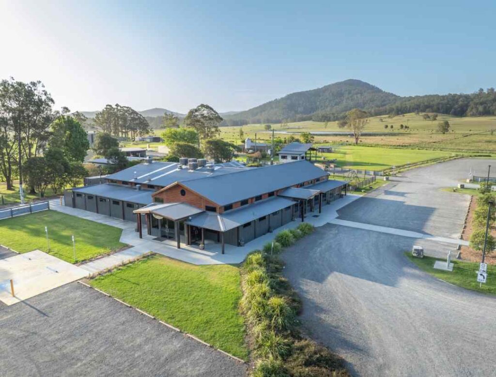 A modern low-rise building with a metal roof on a rural campus, surrounded by green fields and distant hills.