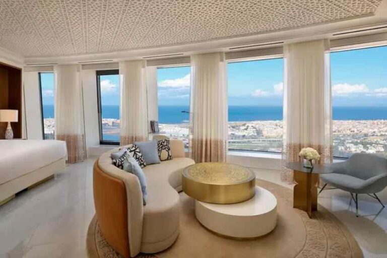 Luxurious hotel suite living area with curved beige sofa, gold coffee table, and large windows overlooking the ocean.