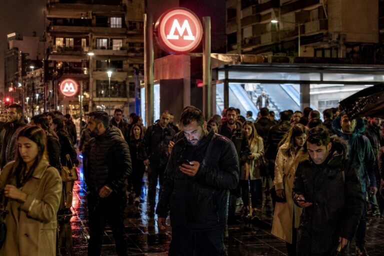 Crowd of pedestrians at a rainy night near a metro station entrance with red circular M signs, reflecting on wet pavement.