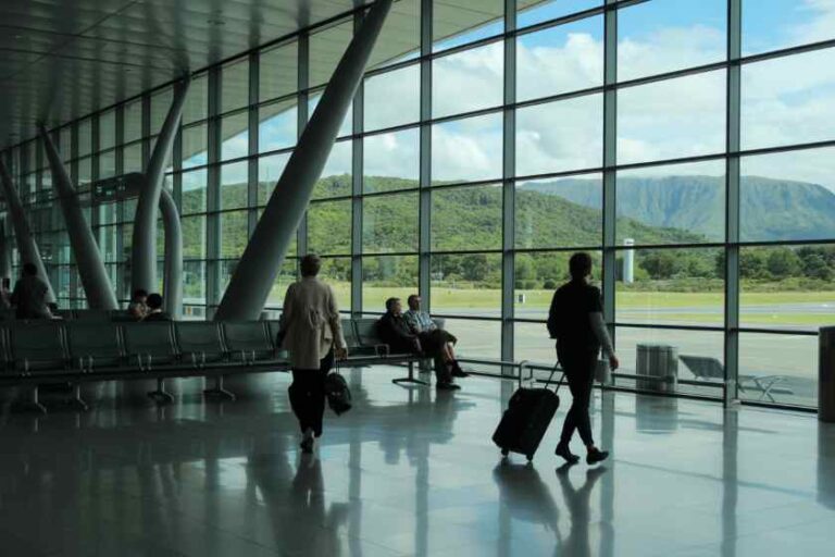 People with rolling suitcases walk through a bright airport terminal with floor-to-ceiling windows and views of a green landscape outside.