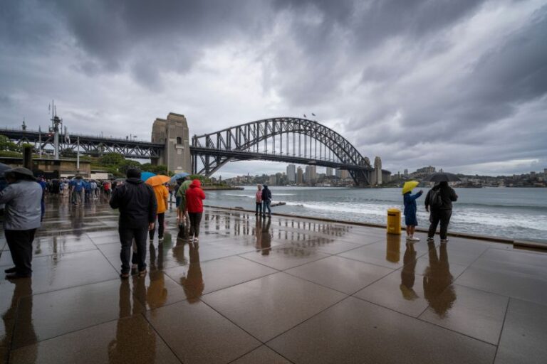 People with umbrellas on a rain-soaked promenade by Sydney Harbour Bridge under a cloudy sky.