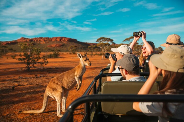 Group of tourists in a safari jeep taking photos as a kangaroo stands by the vehicle in a red desert landscape under a blue sky
