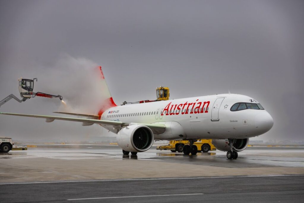 Austrian Airlines jet on the tarmac being de-iced by an elevated spray arm releasing red fluid near the tail area.