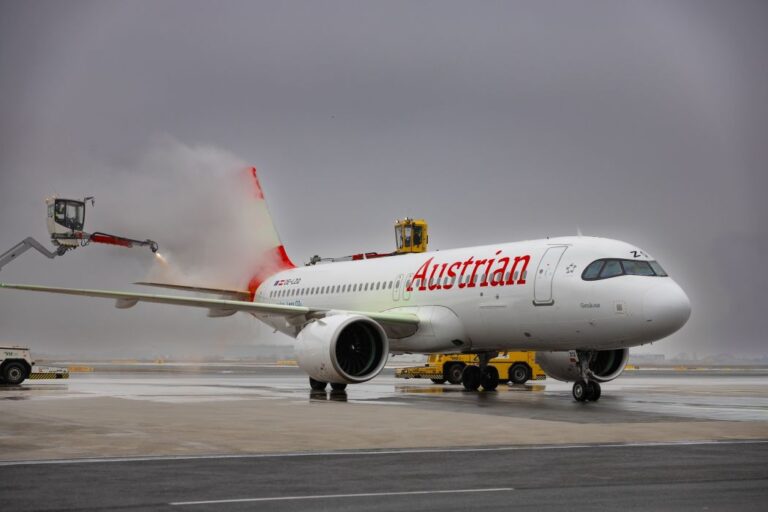 Austrian Airlines jet on the tarmac being de-iced by an elevated spray arm releasing red fluid near the tail area.
