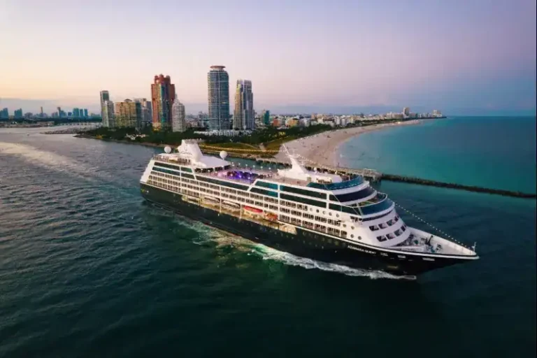 Large white cruise ship sailing along a tropical coastline with a skyline of high-rise buildings in the background at sunset.