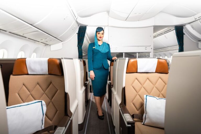 Flight attendant in teal uniform standing in a beige airplane cabin aisle.