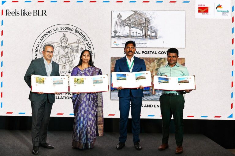 Four professionals stand on a stage holding long white folders with stamps, at a postal event with a 'BLR' backdrop and India Post logos in the background.