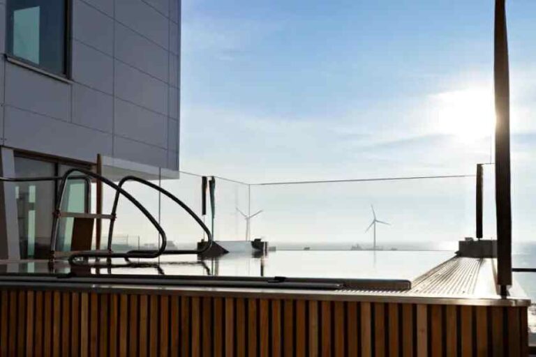 Rooftop hot tub with glass railing, modern building, and distant wind turbines against a bright sunset sky.