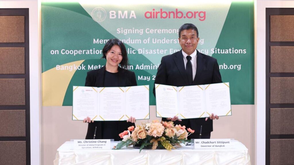 Two professionals in business attire display signed documents at a formal signing ceremony with a green banner in the background.