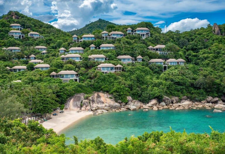 Terraced hillside resort with white-roofed villas among lush greenery overlooking a sandy beach and turquoise bay.